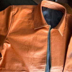 May include: A tan leather jacket featuring a collared neckline and a full-length zip. The jacket has a black lining and visible stitching. The leather has a smooth, slightly glossy finish.