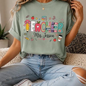 Puede incluir: Una camiseta verde salvia con un gráfico colorido que incluye la palabra TEACHER, donde cada letra está formada por útiles escolares como lápices, libros y crayones. Debajo de la palabra, el texto Mrs. Jessica está impreso en cursiva negra.