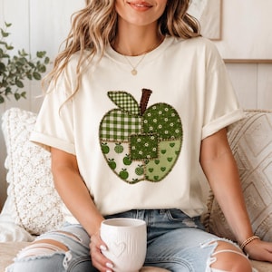 Puede incluir: Una camiseta de manga corta color crema con un gráfico grande de una manzana compuesta por varios patrones de patchwork verdes, incluyendo cuadros vichy, flores y corazones, con un tallo marrón oscuro y un efecto de borde cosido.