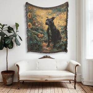 Op de afbeelding: Een geweven wandtapijt met een zwarte hond in een veld met zonnebloemen. Het wandtapijt heeft een gefranjerde rand en hangt aan een witte muur boven een witte bank. Het kleurenpalet omvat geel, groen en zwart.