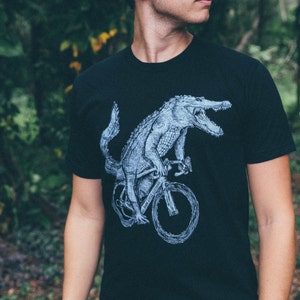 Peut inclure: Un t-shirt noir avec un motif blanc d'un alligator à vélo. L'alligator porte un casque et a une expression déterminée sur son visage.