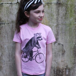 May include: A light pink t-shirt featuring a black and white illustration of a raccoon riding a bicycle. The raccoon is the central focus, with detailed shading. The shirt has short sleeves and a crew neck.