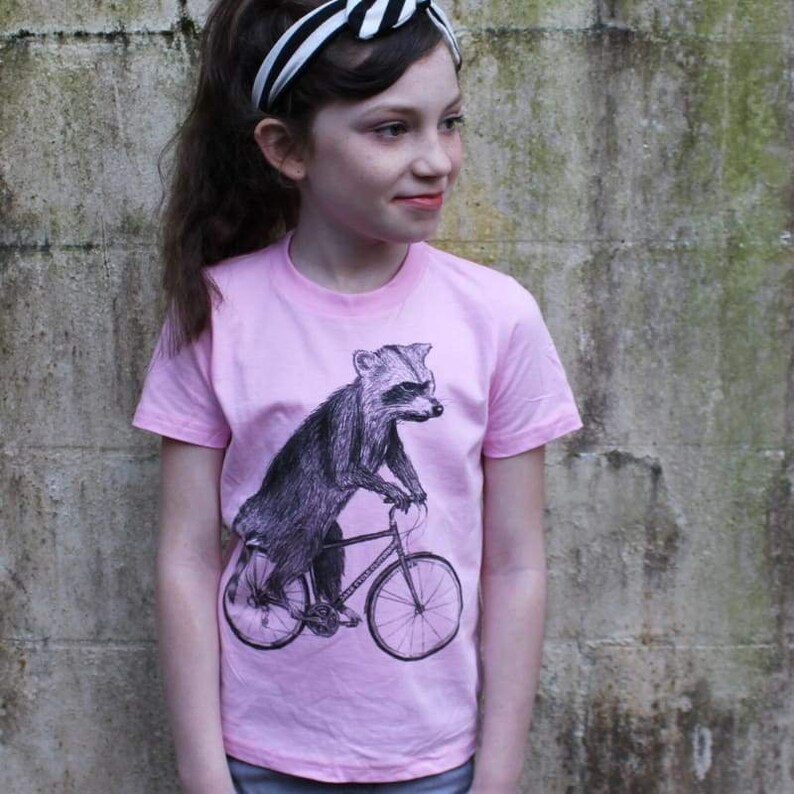 May include: A light pink t-shirt featuring a black and white illustration of a raccoon riding a bicycle. The raccoon is the central focus, with detailed shading. The shirt has short sleeves and a crew neck.