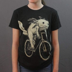 Könnte beinhalten: Schwarzes T-Shirt mit einem weißen Axolotl, das Fahrrad fährt. Der Axolotl hat einen weißen Körper mit schwarzen Augen und einen schwarzen Schwanz. Das Fahrrad hat schwarze Räder und einen schwarzen Rahmen.