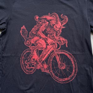 Pode incluir: Uma ilustra&ccedil;&atilde;o vermelha de uma criatura com chifres e um chap&eacute;u de Papai Noel andando de bicicleta. A criatura tem uma l&iacute;ngua longa para fora. O fundo &eacute; preto.