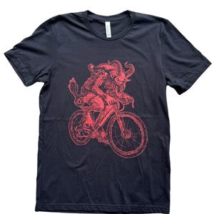 Pode incluir: Uma camiseta preta com uma ilustra&ccedil;&atilde;o vermelha de uma criatura andando de bicicleta. A criatura tem chifres, uma cauda e um chap&eacute;u pontudo.