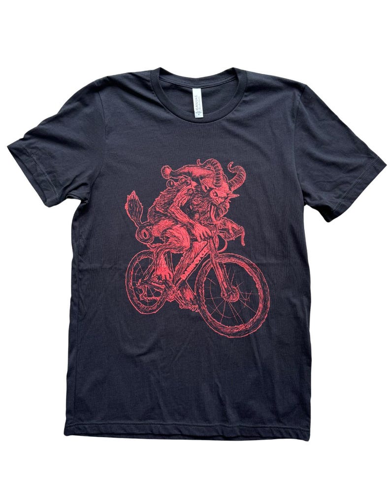 Pode incluir: Uma camiseta preta com uma ilustra&ccedil;&atilde;o vermelha de uma criatura andando de bicicleta. A criatura tem chifres, uma cauda e um chap&eacute;u pontudo.