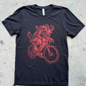 Pode incluir: Camiseta preta com uma ilustra&ccedil;&atilde;o vermelha de uma criatura com chifres andando de bicicleta.