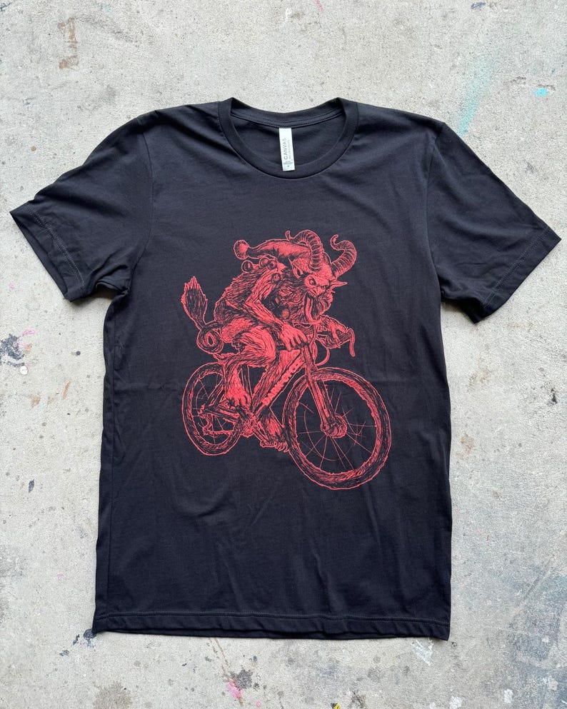 Pode incluir: Camiseta preta com uma ilustra&ccedil;&atilde;o vermelha de uma criatura com chifres andando de bicicleta.