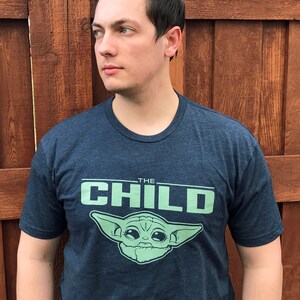 May include: Navy blue t-shirt featuring the text "THE CHILD" in light green, along with an illustration of a character. The shirt is made of a soft material and has short sleeves.