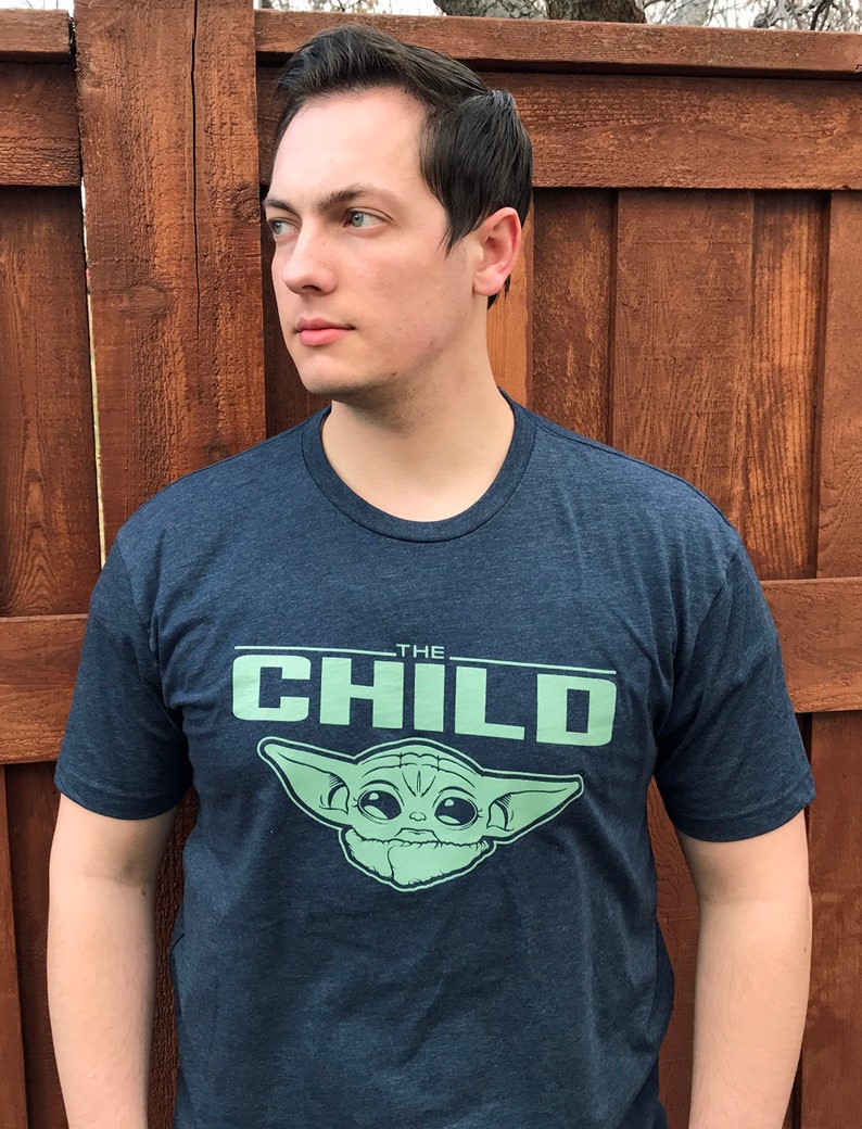 May include: Navy blue t-shirt featuring the text "THE CHILD" in light green, along with an illustration of a character. The shirt is made of a soft material and has short sleeves.
