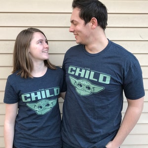 May include: Two people wearing matching navy blue t-shirts with the text "THE CHILD" and a graphic of a character. The shirts are short-sleeved and have a light green design. The people are smiling and looking at each other.