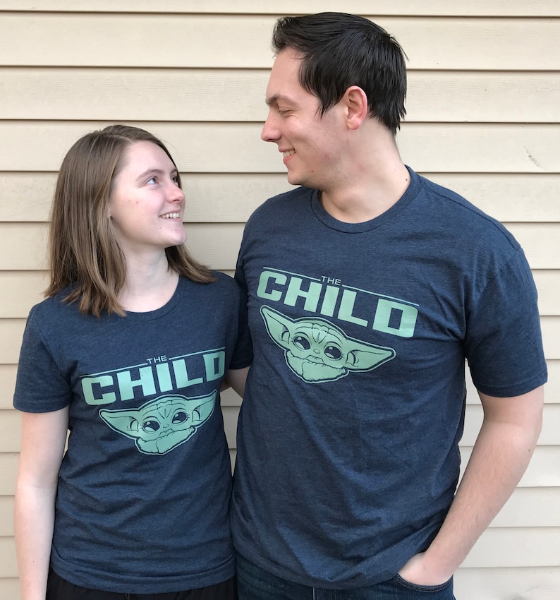 May include: Two people wearing matching navy blue t-shirts with the text "THE CHILD" and a graphic of a character. The shirts are short-sleeved and have a light green design. The people are smiling and looking at each other.