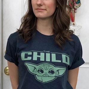 May include: A navy blue t-shirt featuring the text "THE CHILD" above a green graphic of a character from the Star Wars series. The shirt is paired with light blue jeans and a black belt.