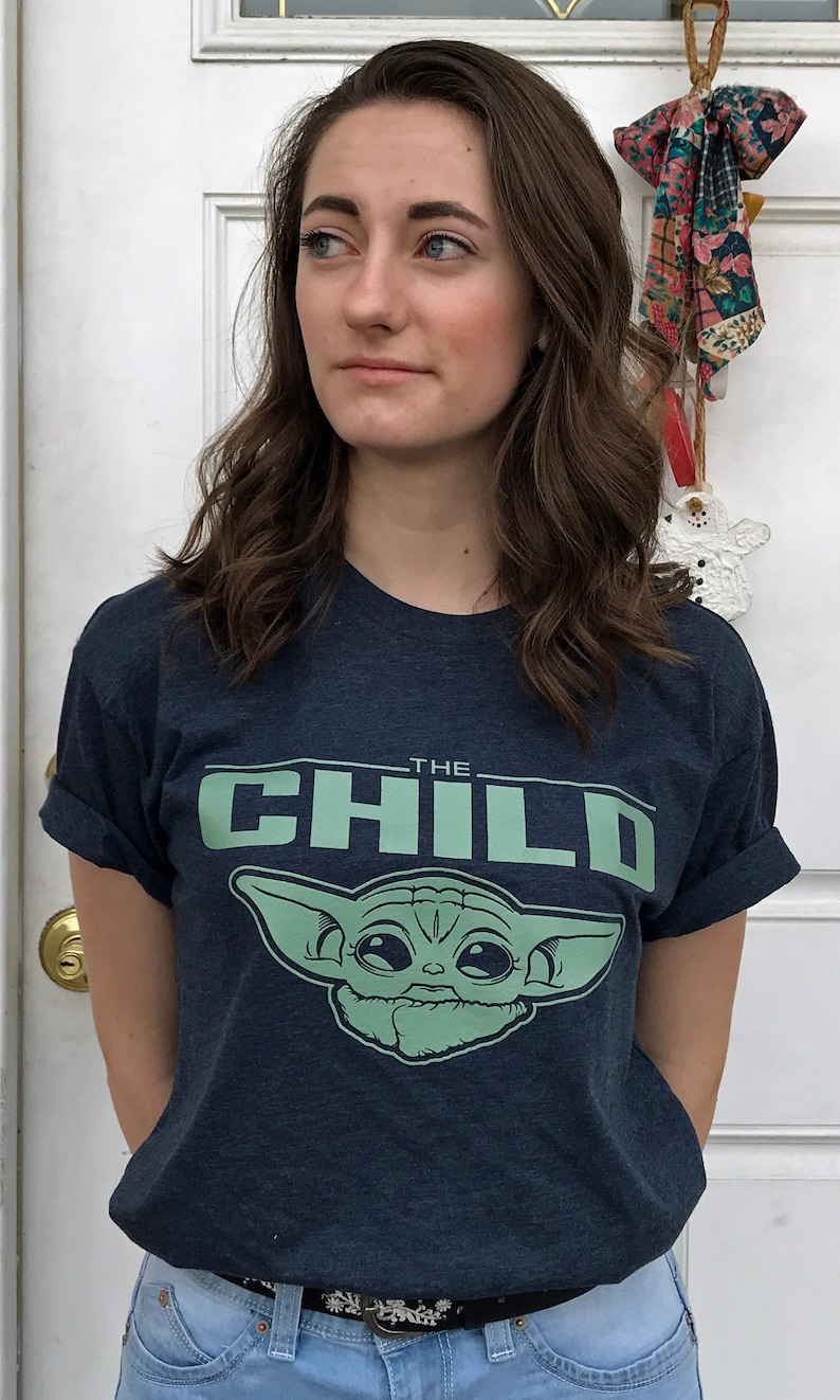 May include: A navy blue t-shirt featuring the text "THE CHILD" above a green graphic of a character from the Star Wars series. The shirt is paired with light blue jeans and a black belt.