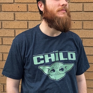 May include: Navy blue t-shirt featuring the text "THE CHILD" above a light green image of Grogu from The Mandalorian. The shirt is short-sleeved and made of a soft material.