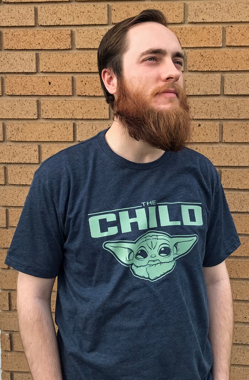 May include: Navy blue t-shirt featuring the text "THE CHILD" above a light green image of Grogu from The Mandalorian. The shirt is short-sleeved and made of a soft material.