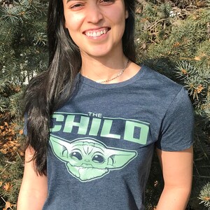May include: A navy blue t-shirt featuring the text "THE CHILD" in green, along with a graphic of the character from the Star Wars series, The Mandalorian. The shirt is paired with blue jeans.