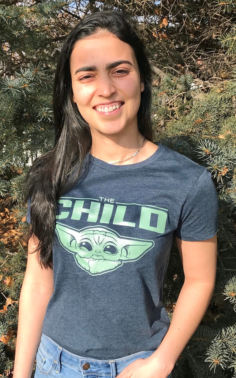 May include: A navy blue t-shirt featuring the text "THE CHILD" in green, along with a graphic of the character from the Star Wars series, The Mandalorian. The shirt is paired with blue jeans.