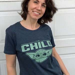 May include: Navy blue t-shirt featuring the text "THE CHILD" above a light green graphic of a character. The shirt is a crew neck style and appears to be made of a soft, comfortable material.