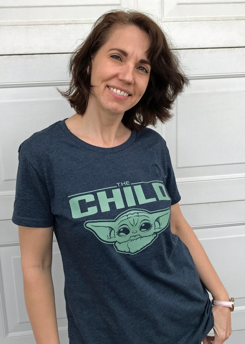 May include: Navy blue t-shirt featuring the text "THE CHILD" above a light green graphic of a character. The shirt is a crew neck style and appears to be made of a soft, comfortable material.