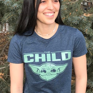 May include: A navy blue t-shirt featuring the text "THE CHILD" above a green graphic of Grogu from The Mandalorian. The shirt is worn by a person with long black hair and blue jeans.