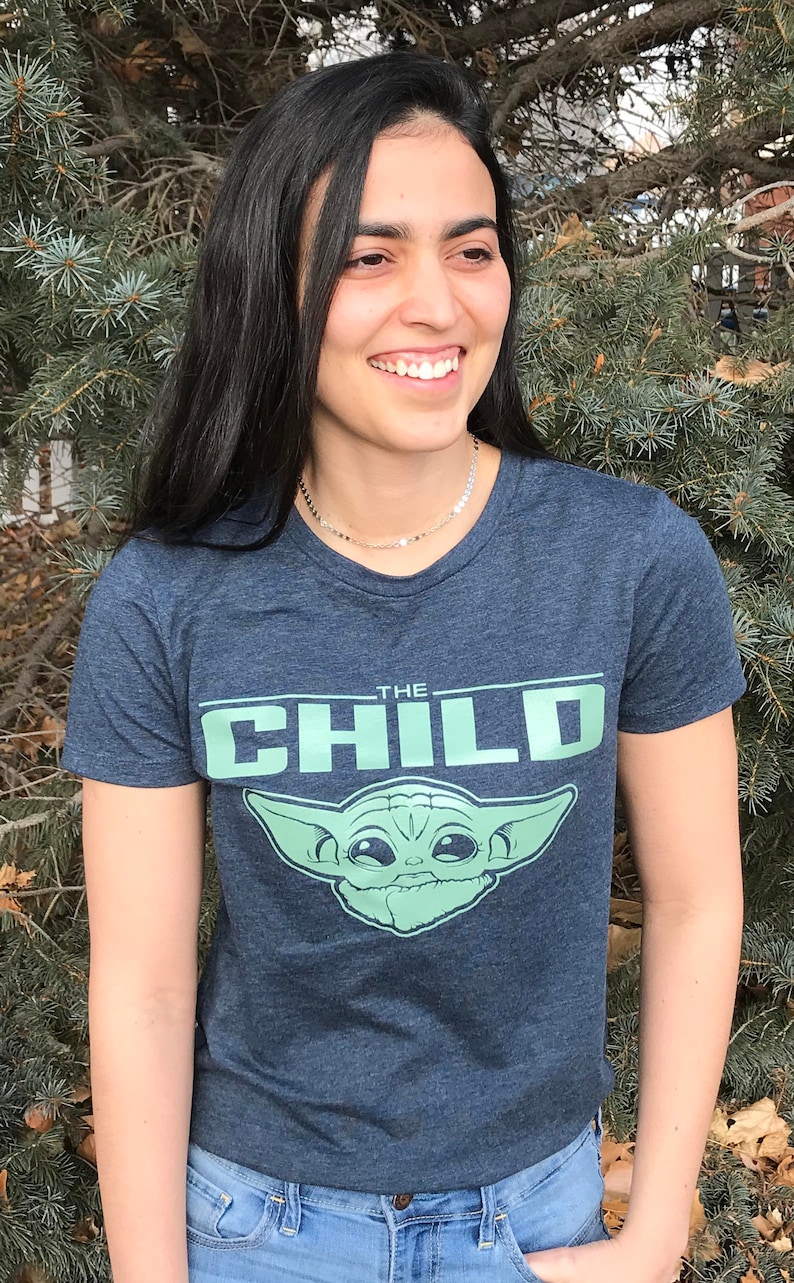 May include: A navy blue t-shirt featuring the text "THE CHILD" above a green graphic of Grogu from The Mandalorian. The shirt is worn by a person with long black hair and blue jeans.
