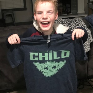 May include: Navy blue t-shirt featuring a green graphic of a character with large ears and the text "THE CHILD". The shirt is held up by a person. The person is smiling and laughing.
