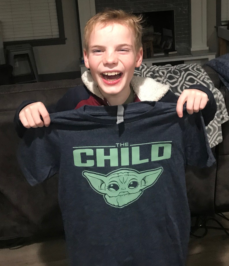 May include: Navy blue t-shirt featuring a green graphic of a character with large ears and the text "THE CHILD". The shirt is held up by a person. The person is smiling and laughing.