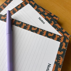 May include: Lined notepads with a tiger print border and the word "Notes". A lavender pen lies across the notepads. The background is a light brown wooden surface. The notepads are stacked.