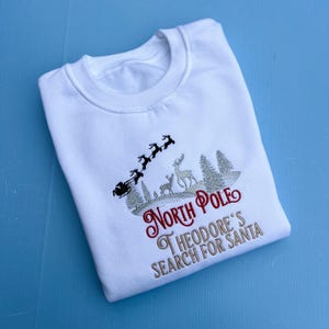 May include: White sweatshirt with an embroidered design of Santa's sleigh, reindeer, and trees. The text "North Pole" and "Theodore's Search for Santa" are in red and grey thread.