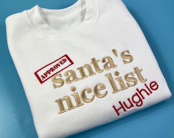 May include: White sweatshirt with gold embroidery that reads "Santa's nice list" and a red stamp that says "APPROVED". The name "Hughie" is embroidered in red below the text.