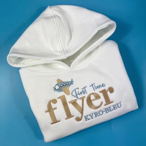 Könnte beinhalten: Weißer Kapuzenpullover mit einem blauen Flugzeug und goldener Stickerei mit der Aufschrift "First Time Flyer KYRO-BLEU".