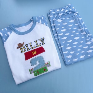Puede incluir: Un conjunto de pijama blanco y azul con el nombre "BILLY" y el número "2" bordados en la camisa. La camisa tiene mangas azules y un cuello azul. Los pantalones de pijama son de color azul claro con un estampado de nubes blancas.