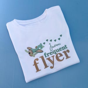 以下が含まれることがあります： 緑の飛行機と「Jaxon frequent flyer」という茶色の文字が書かれた白いTシャツです。
