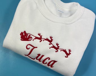 May include: White Christmas sweatshirt with red embroidered Santa Claus and reindeer pulling a sleigh. The name "Luca" is embroidered below the image.