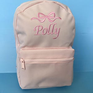 May include: A pale pink backpack with a front zipped pocket. The backpack has a pink embroidered bow and the name "Polly" in pink script. It features a top handle and adjustable shoulder straps.