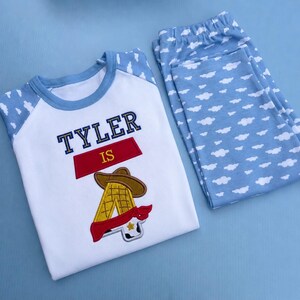 Può includere: Una maglietta bianca con maniche raglan blu e pantaloni abbinati, entrambi caratterizzati da un motivo a nuvole bianche su sfondo azzurro. La maglietta mostra il testo ricamato TYLER IS sopra un grande numero 4 con cappello da cowboy.