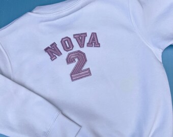 May include: White sweatshirt with the word "NOVA" and the number "2" embroidered in a light purple color on the back. The sweatshirt has a crew neck and long sleeves.