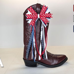 May include: A burgundy cowboy boot adorned with a patriotic bow featuring red and white polka dots, white and blue ribbons. The boot has a pointed toe and a black heel. The bow and ribbons add a festive touch to the boot.