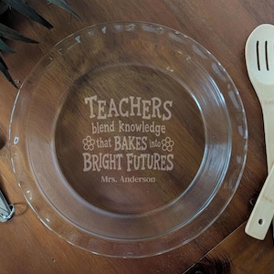 May include: A clear glass pie dish with etched text that reads "Teachers blend knowledge that BAKES into BRIGHT FUTURES Mrs. Anderson." Two wooden cooking utensils and a set of measuring spoons are also visible.