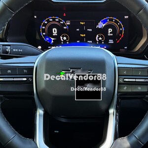 May include: A black steering wheel with a carbon fiber trim and a black decal that says "Decalvendor88". The steering wheel has a silver air bag cover and a silver button on the left side.