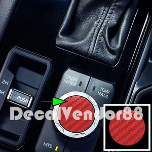 May include: Red carbon fiber decal for a car's drive mode selector knob. The decal has a silver ring around the edge. The text "DRIVE MODE" is visible on the knob. The decal is shown next to a black and silver knob with the text "TOW HAUL".