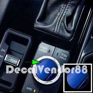 May include: Close-up of a car's center console with a blue knob labeled "Drive Mode". The knob has a silver ring with markings for different drive modes, including "Eco", "Sport", and "Snow". The console also has buttons for "2H", "4H", "Push", "Mts", and "Tow Haul".