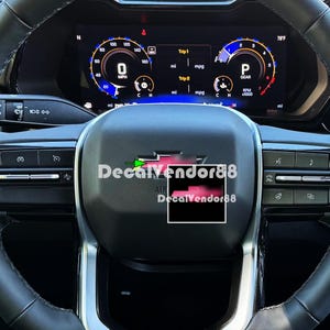 May include: A black leather steering wheel with a silver trim and a red and black decal that says "DecalVendor88". The steering wheel is in a car with a digital dashboard showing the speed, RPM, and other information.