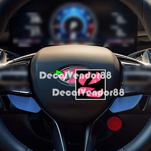 May include: A black car steering wheel featuring a central emblem with a green arrow pointing toward it. The steering wheel includes blue paddle shifters on the sides, a red circular button on the lower right, and the text DecalVendor88 overlaid twice.