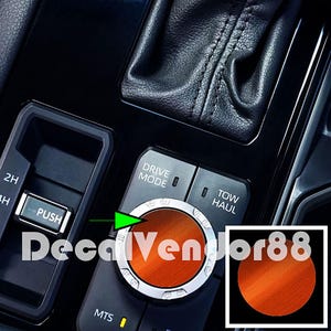 May include: Close-up of a car's drive mode selector knob with a brushed orange finish. The knob is labeled "Drive Mode" and has a silver ring around the edge. The knob is surrounded by other buttons, including "2H", "4H", "Push", "Mts", and "Tow Haul".