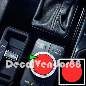 May include: Close-up of a car's drive mode selector knob with a red button. The knob has a silver ring with markings for different drive modes, including 2H, 4H, and MTS. The text "Drive Mode" is visible above the knob. The knob is surrounded by a black plastic panel with the text "Tow Haul" to the right.