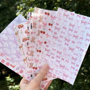 Op de afbeelding: Een set van zes decoratieve papieren vellen met verschillende patronen. De patronen omvatten roze strikken, kersen en beren. De vellen zijn wit met roze en rode accenten.