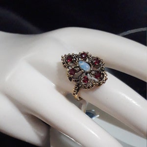 Puede incluir: Un anillo de estilo vintage con un diseño floral. El anillo presenta una piedra central ovalada de color azul claro, rodeada de piedras rojas redondas más pequeñas. El metal del anillo tiene un tono dorado con intrincados detalles de filigrana.